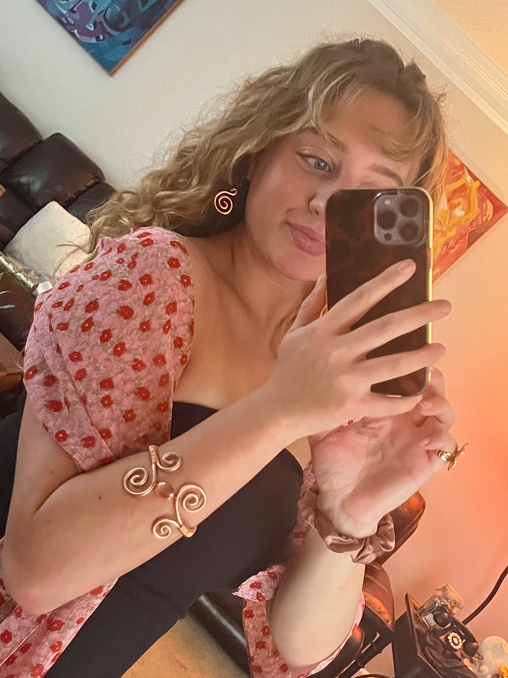 Woman taking a mirror selfie wearing a floral top and gold jewelry.