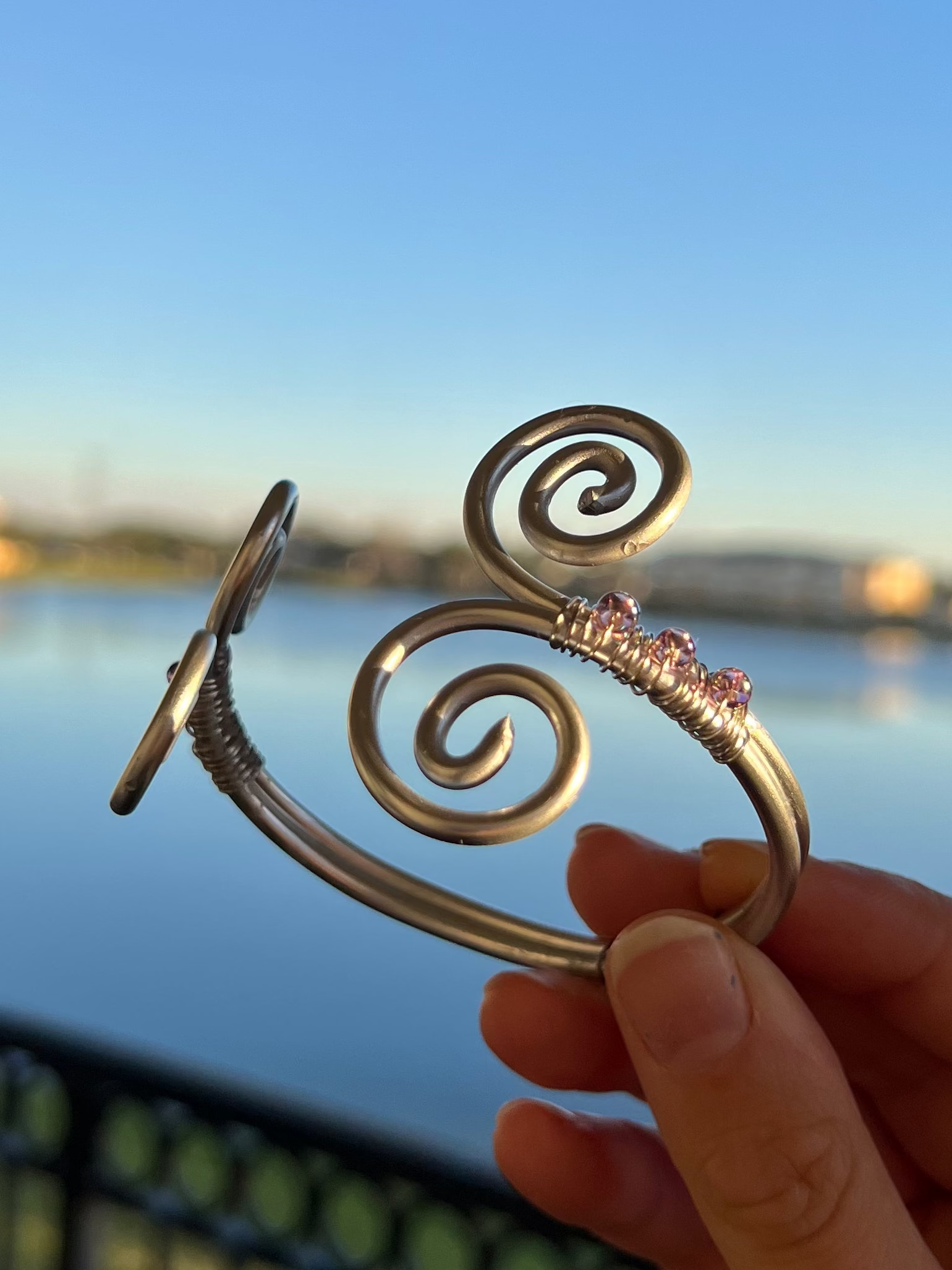 Hand holding a decorative metal object with a spiral design against a scenic background of water and sky.