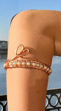 Person wearing a gold bracelet with a heart design against a blue sky and water background