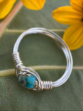 Silver wire-wrapped ring with a blue stone on a green leaf background with yellow flowers.