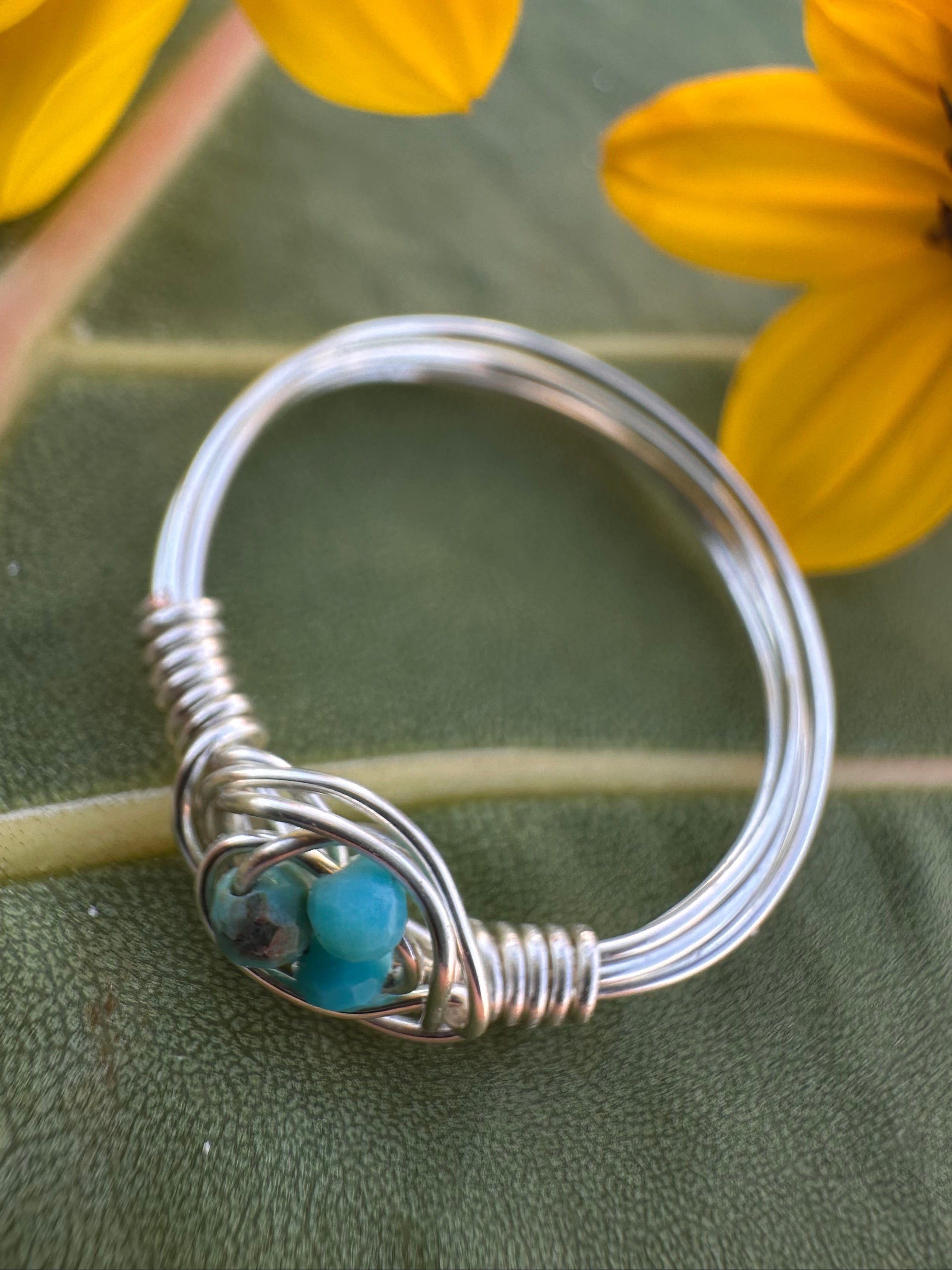 Silver wire-wrapped ring with a blue stone on a green leaf background with yellow flowers.