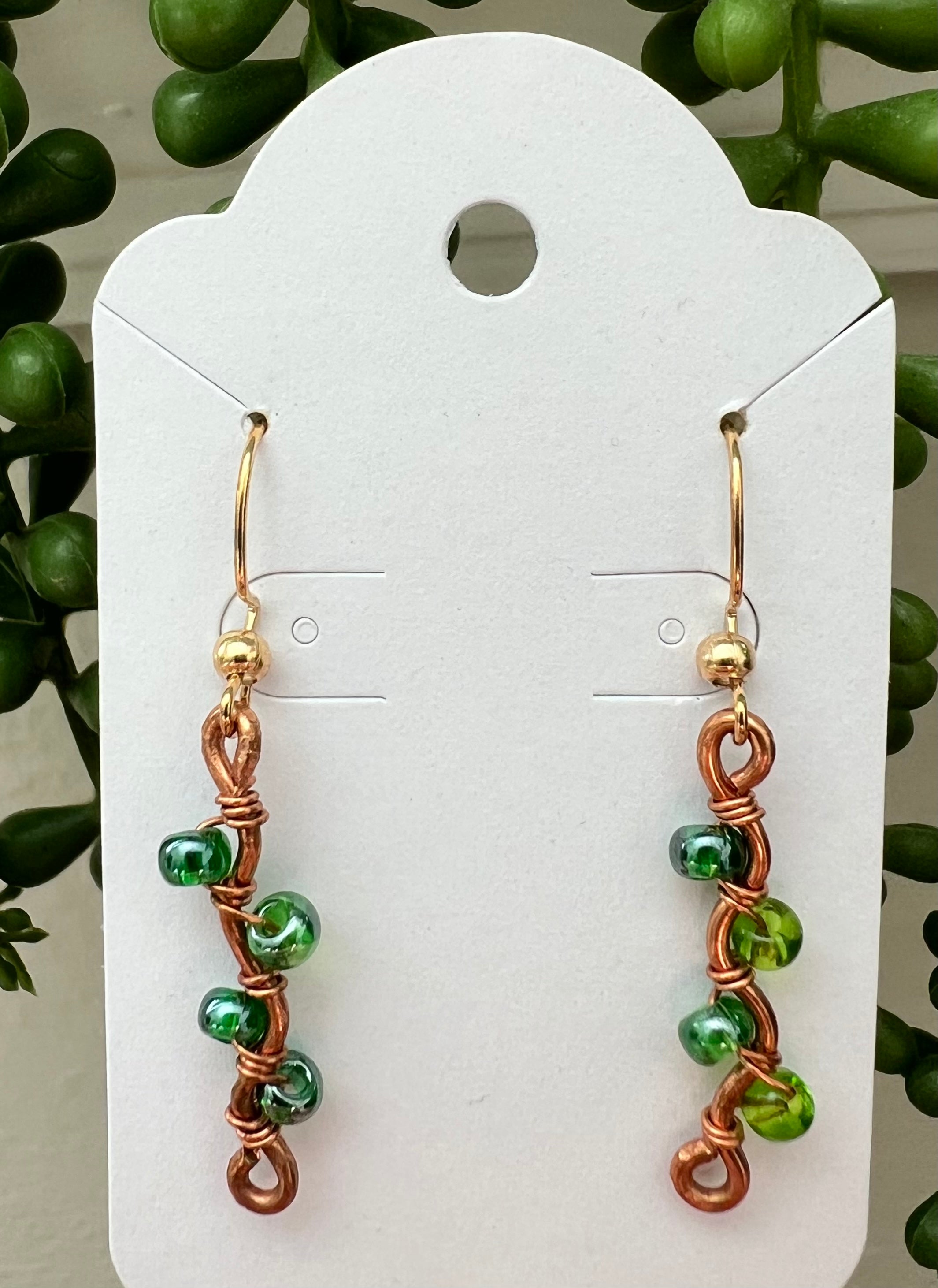 Pair of green and copper wire-wrapped earrings on a white card with a green leafy background.