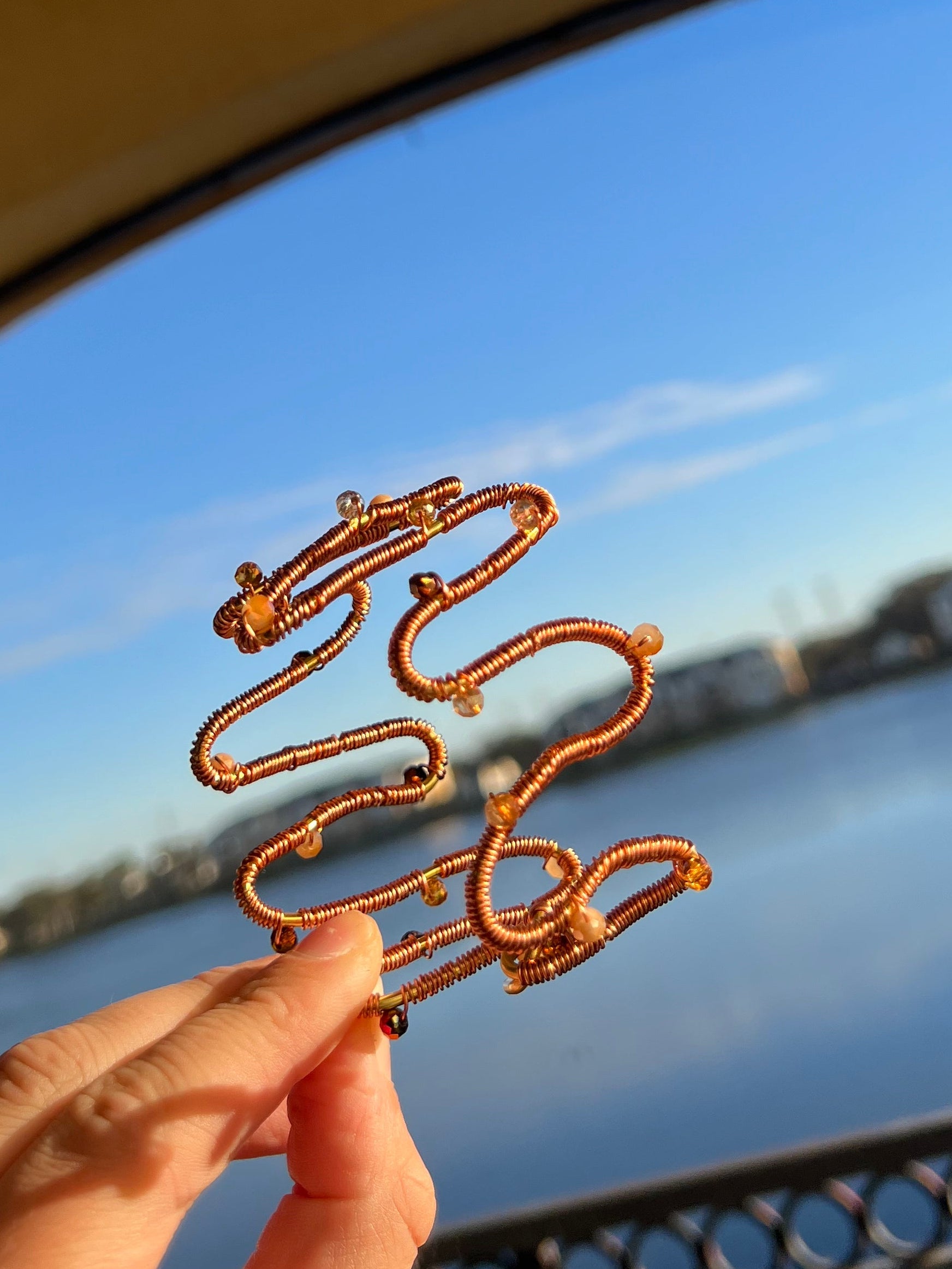 Gold wire earrings held in front of a window with blue sky and building outside.
